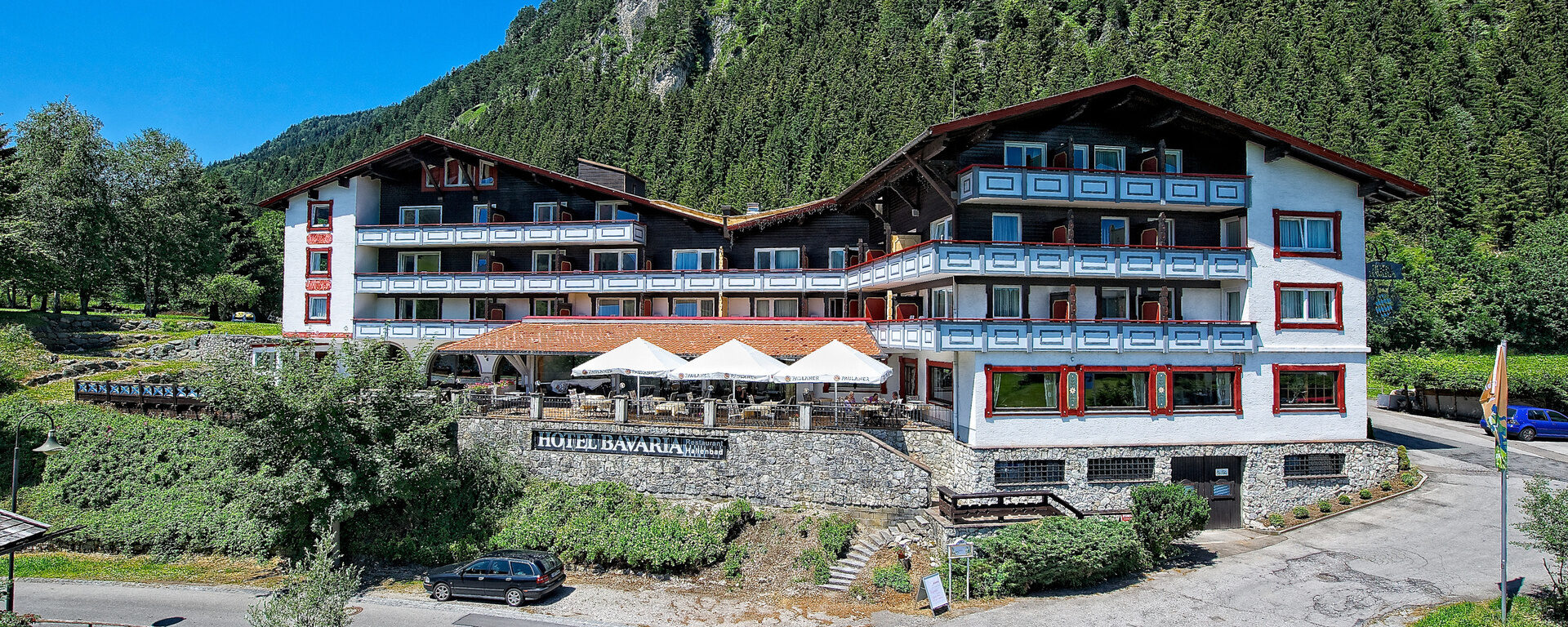Das wei&szlig;-rote Geb&auml;ude des Hotels Bavaria Pfronten in einer Alpenlandschaft.